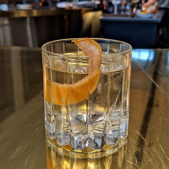 A highball glass with a clear cocktail, ice, and an orange rind