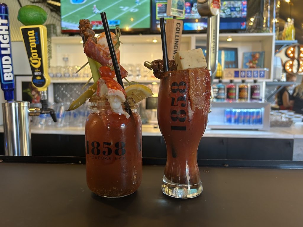 two epic caesars from 1858 caesar bar sitting on the bar and garnished with bacon, shrimp, havarti and lobster tail