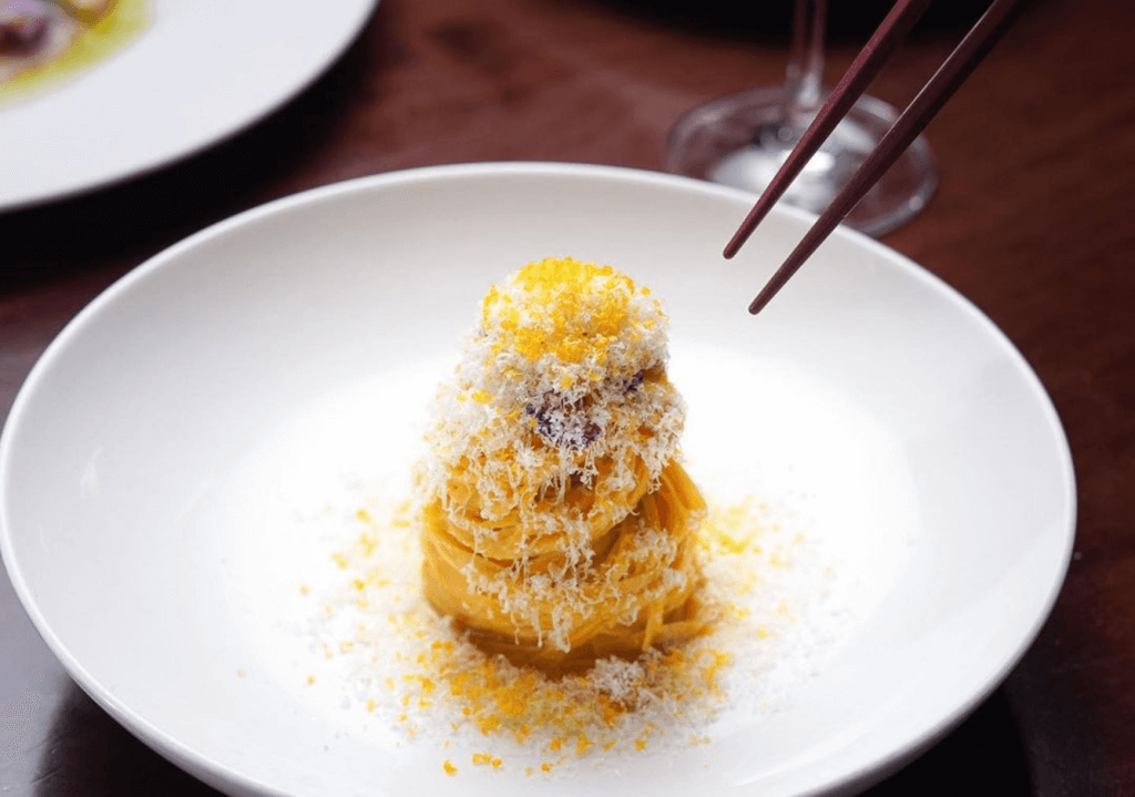 A small nest of handcrafted pasta with orange zest and cheese grated on top, sitting on a white plate with tweezers set to adjust