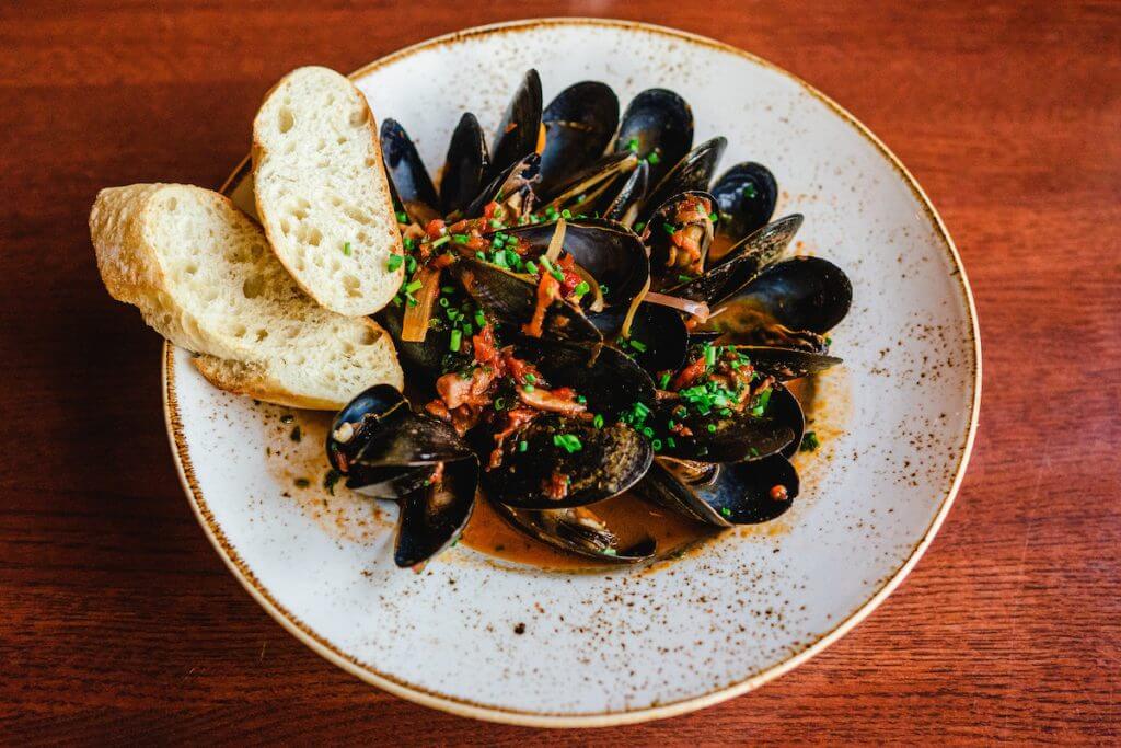 mussels and two slices of baguette in a red sauce in a shallow white speckled bowl