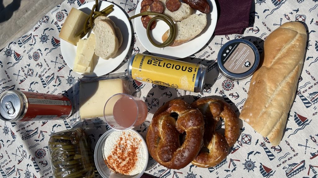 Overhead shot of the picnic basket contesnts from farm 2 door spread out on a blanket including a baguette, can of honey drink, two pretzels, cured meat, cheese and more