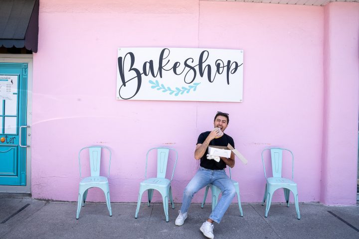 zain sitting on turquoise chair outside of wannamaker's bakery eating a donut in front of a pink wall and bakeshop sign