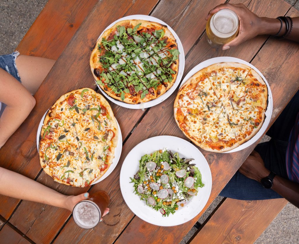 overhead shot of three pizzas and a salad at fixed gear with Gabby and Royden's arms each holding a beer.