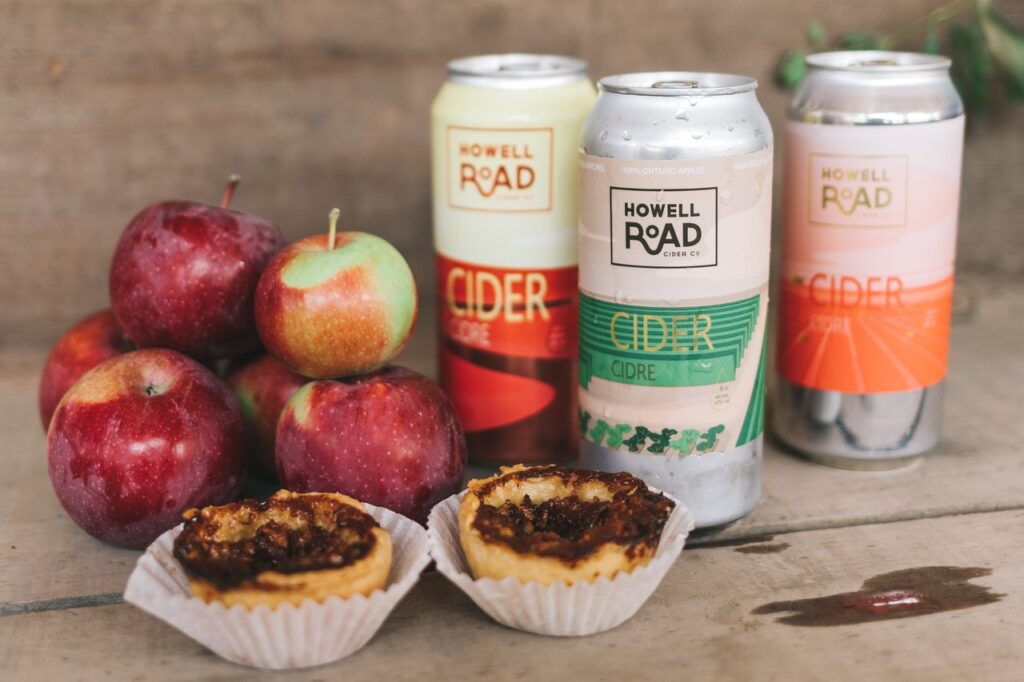 Photo of apples stacked beside three cans of howell road cider and two apple pie tarts