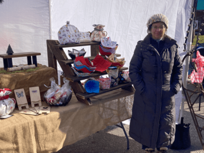 Woman standing in front of table displaying craft items at an outdoor market.