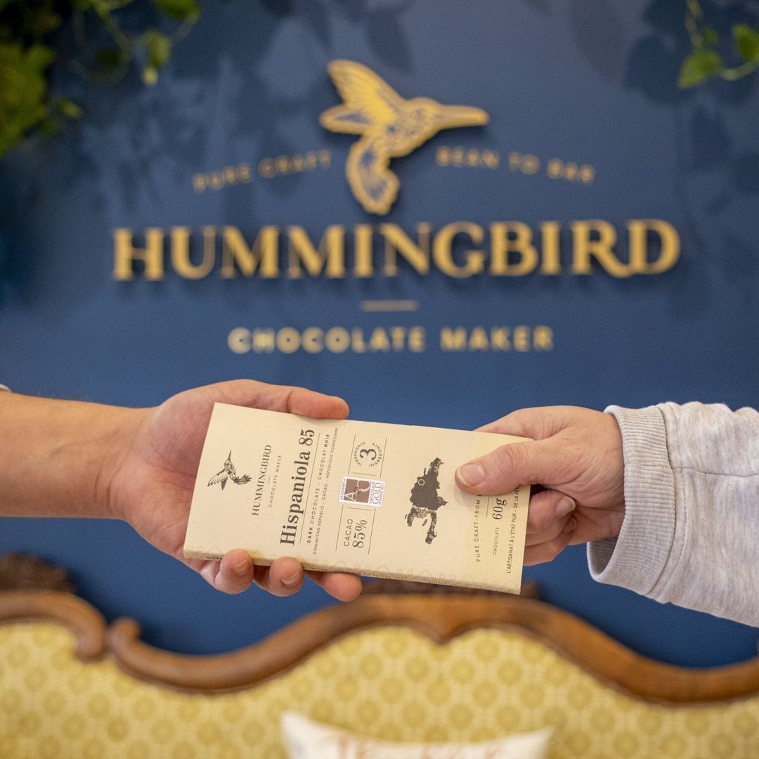 two hands holding a hummingbird chocolate bar in front of the Hummingbird Chocolate sign