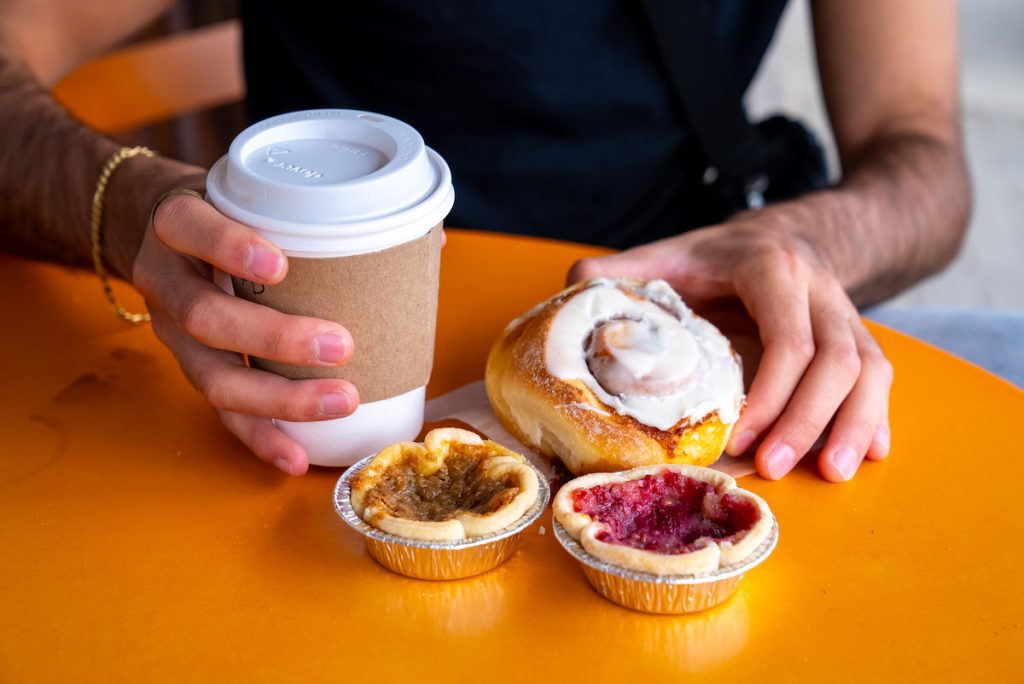 close up of zain holding coffee and cinnamon bun as well as two tarts on an orange table