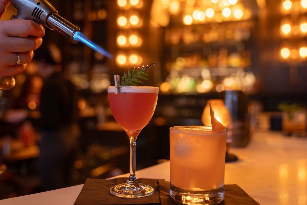 two cocktails at barkeep, one pink one with a cedar spring being singed by a brulee torch and the other shorter and orange with an orange peel twist garnish
