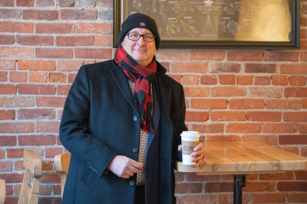 Paul holding a coffee and leaning against a table at St. Paul Roastery with a coffee