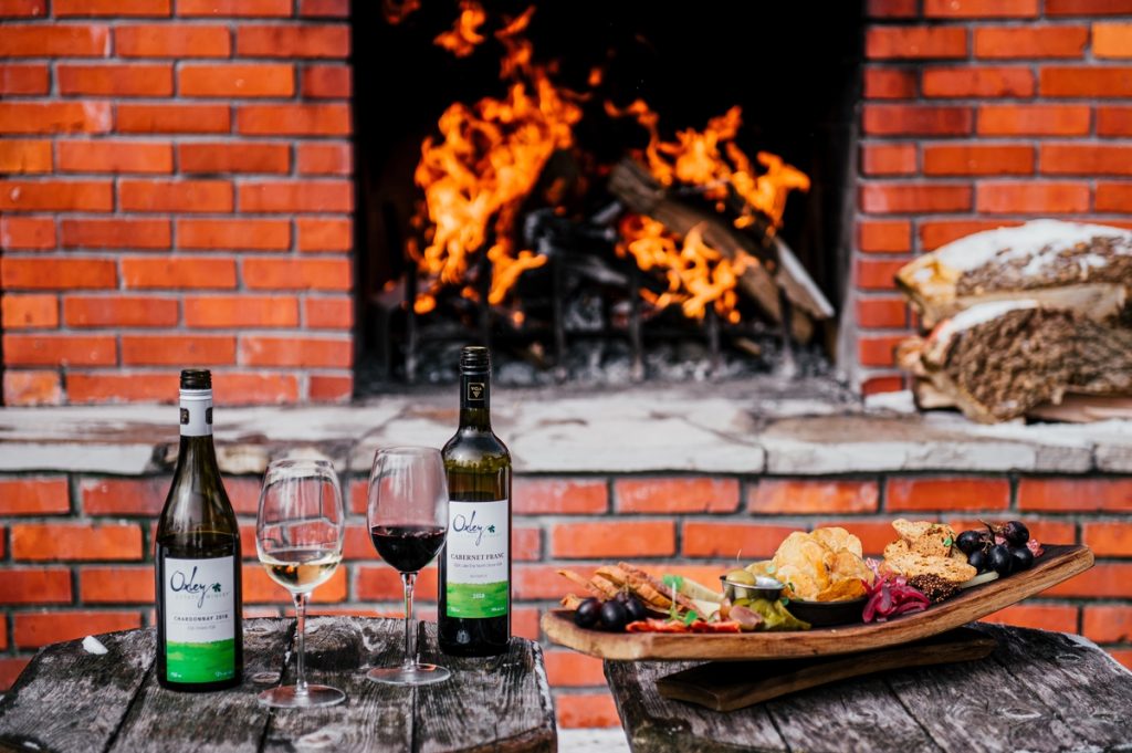 two bottles of wine, two glasses of wine and a charcuterie board set against the backdrop of an open air hearth with a raging fire