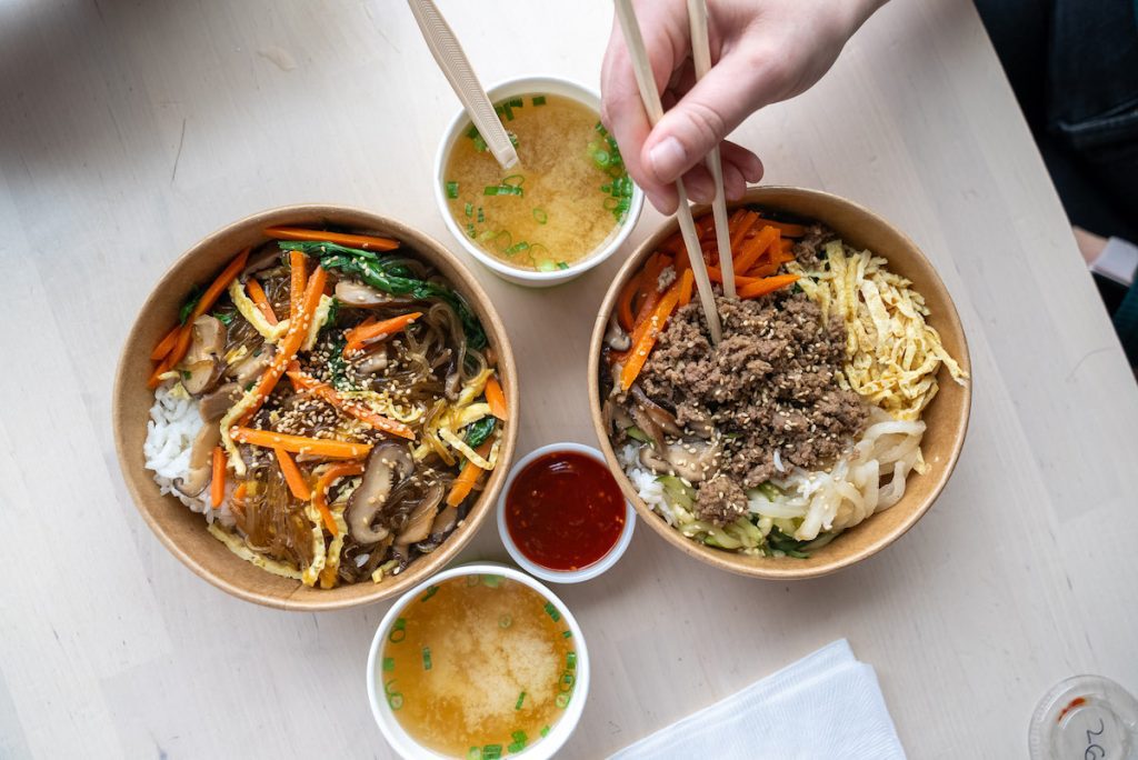An overhead shot of bipimbap, japchae and two miso soups plus hot sauce from BopShop. The bipimbap has a hand holding two chopsticks in it.