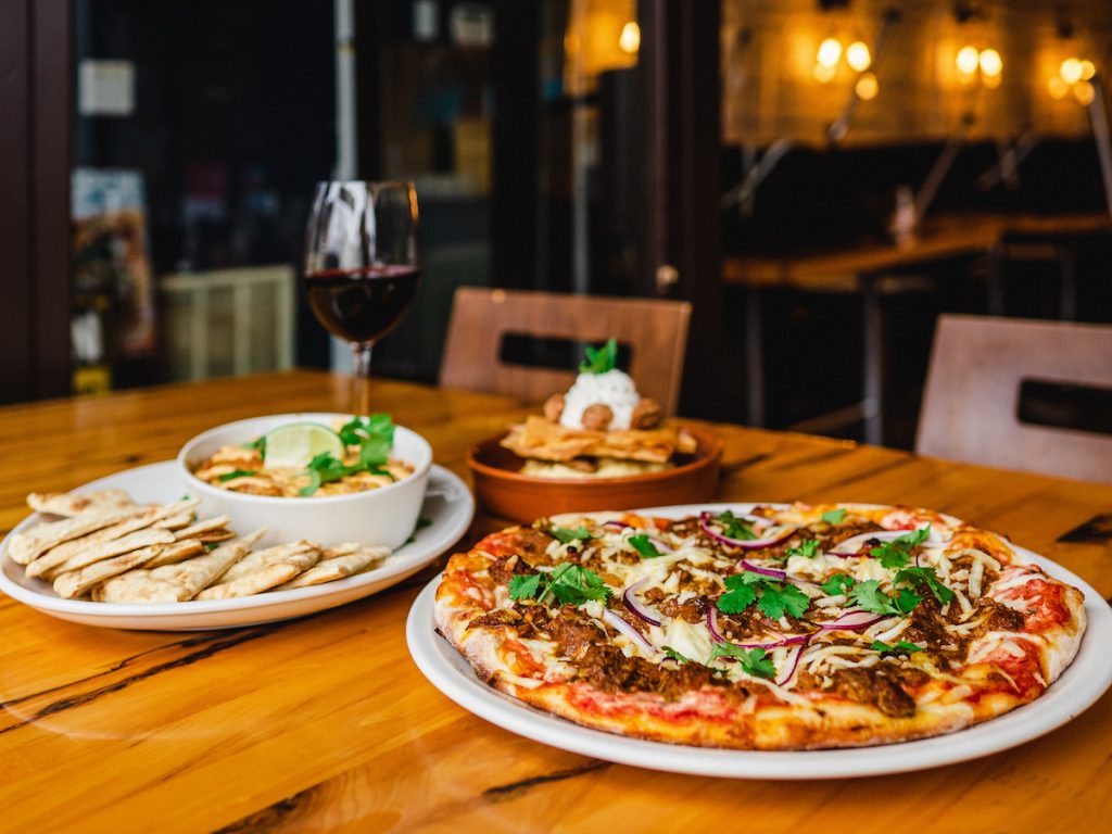 a pizza, hummus and some pita bread with a glass of red wine on the table at atomica