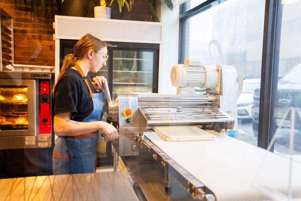 Emily rolling out dough on machine at Swell Bakery in front window