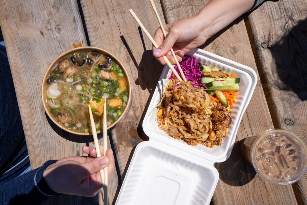 Photo of two hands eating with chopsticks. One from a pad tai dish and a second from a pho noodle soup dish