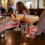 Image of two cocktail glasses clinking in a dark setting over a table.