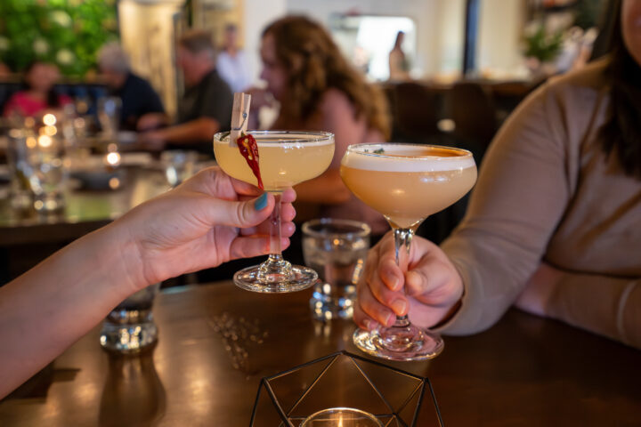 Image of two cocktail glasses clinking in a dark setting over a table.