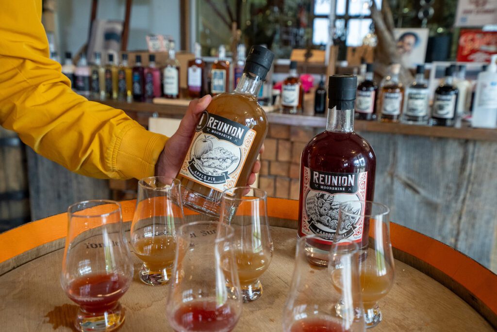 Zain holding a bottle of moonshine behind glasses of it at Top Shelf Distillers