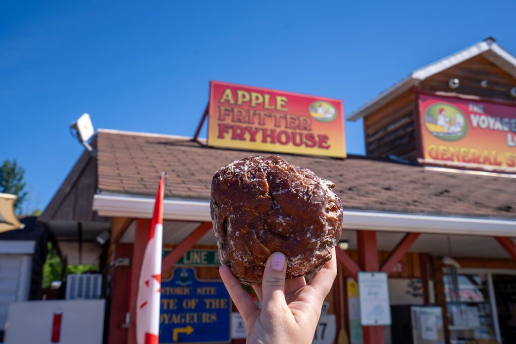 apple fritter held up to Voyageurs' Lodge sign