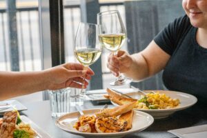 Two glasses of wine clicking with plates of pasta underneath on table