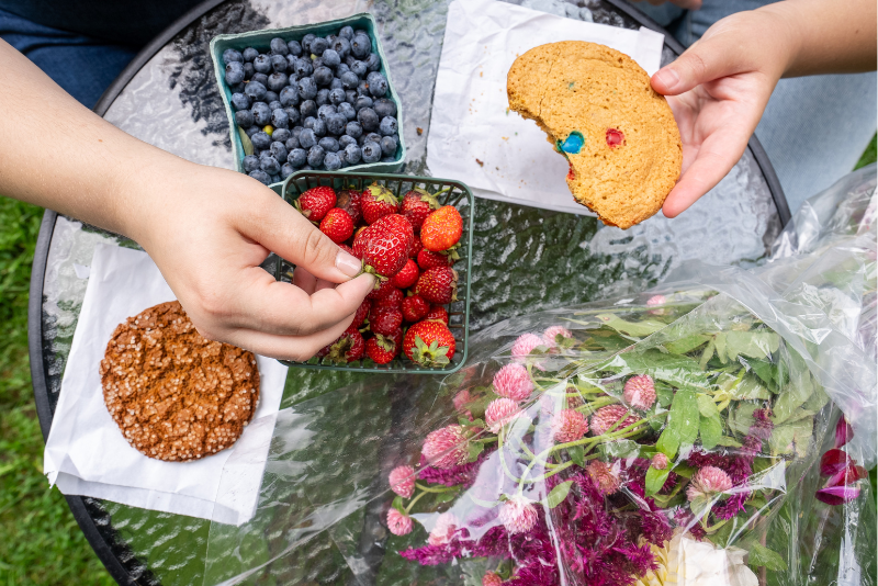 Over head shot of two hands with cookies, a strawberry basket, and bouquet of flowers