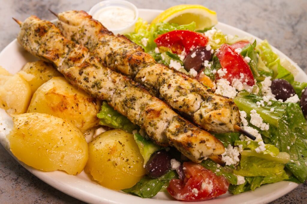 Close up of plate of greek inspired food with chicken skewers, salad, potatoes, and lemon wedges.