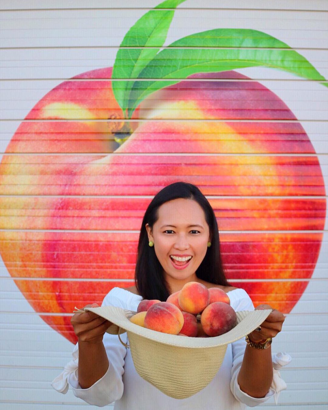 Woman holding hat pull of peaches standing in front of a wall with mural of peach painted.